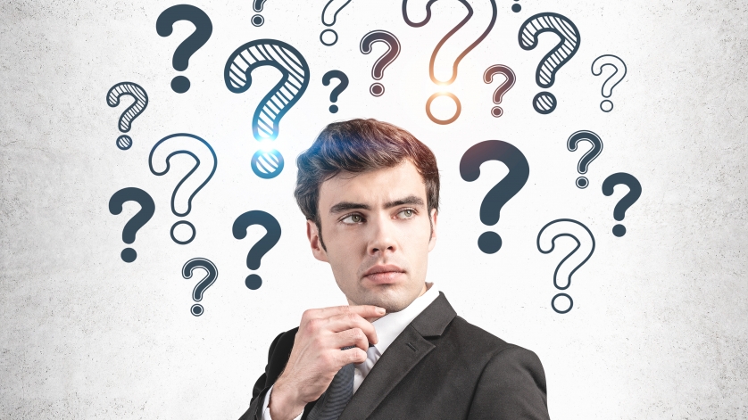 Thoughtful young businessman in suit standing near concrete wall with question marks drawn on it.