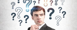 Thoughtful young businessman in suit standing near concrete wall with question marks drawn on it.